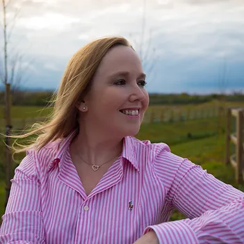 Lisa Browning, owner of Midlands Stress Management is outdoors with an arm resting on a fence or gate. Shes looking happily to her left with a smile on her face and the wind gently catching her hair.