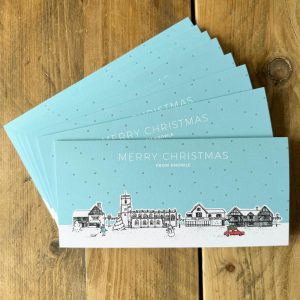 Eight Christmas Cards fanned on wooden table. The image featues line drawings of Knowle buildings at Christmas with a blue sky