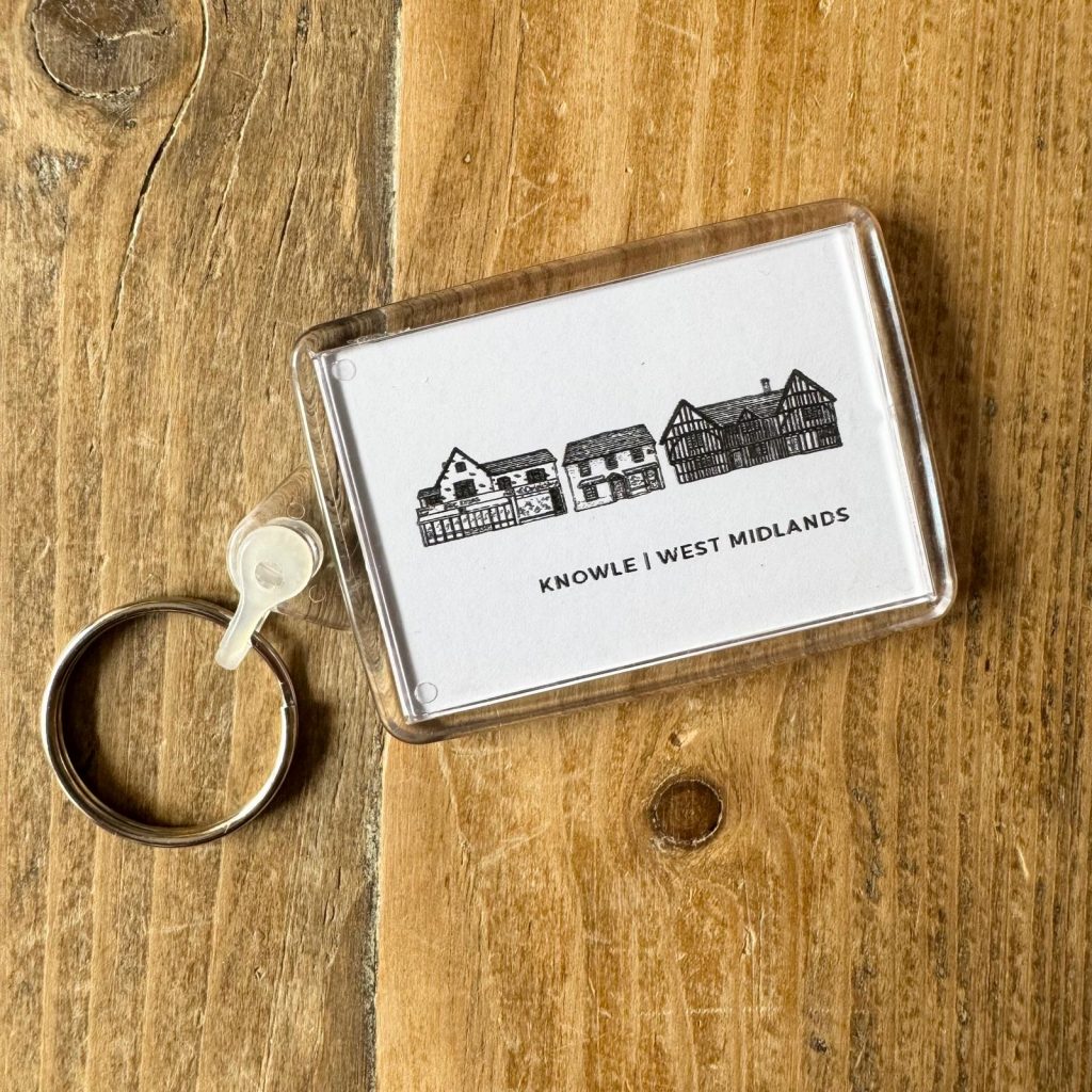 A white keyring with black lne drawings of distinctive buildings in Knowle. The keyring sits on a wooden table.