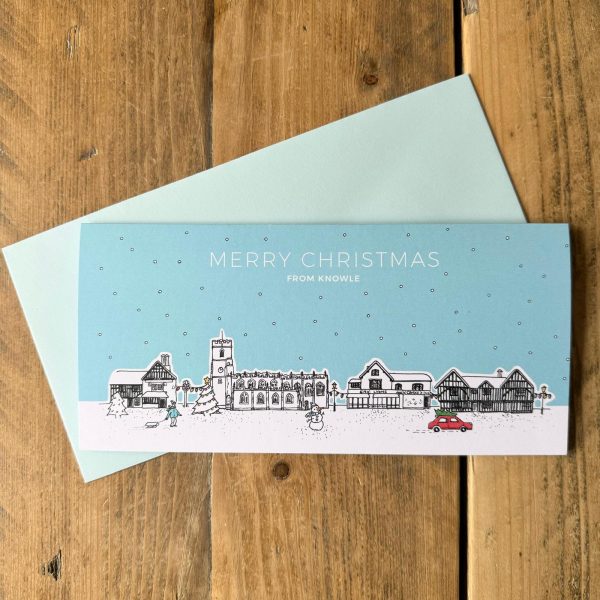 A Christmas card laying on an envelope on a wooden table. The card design is a Christmas scene featuring prominent buildings of Knowle