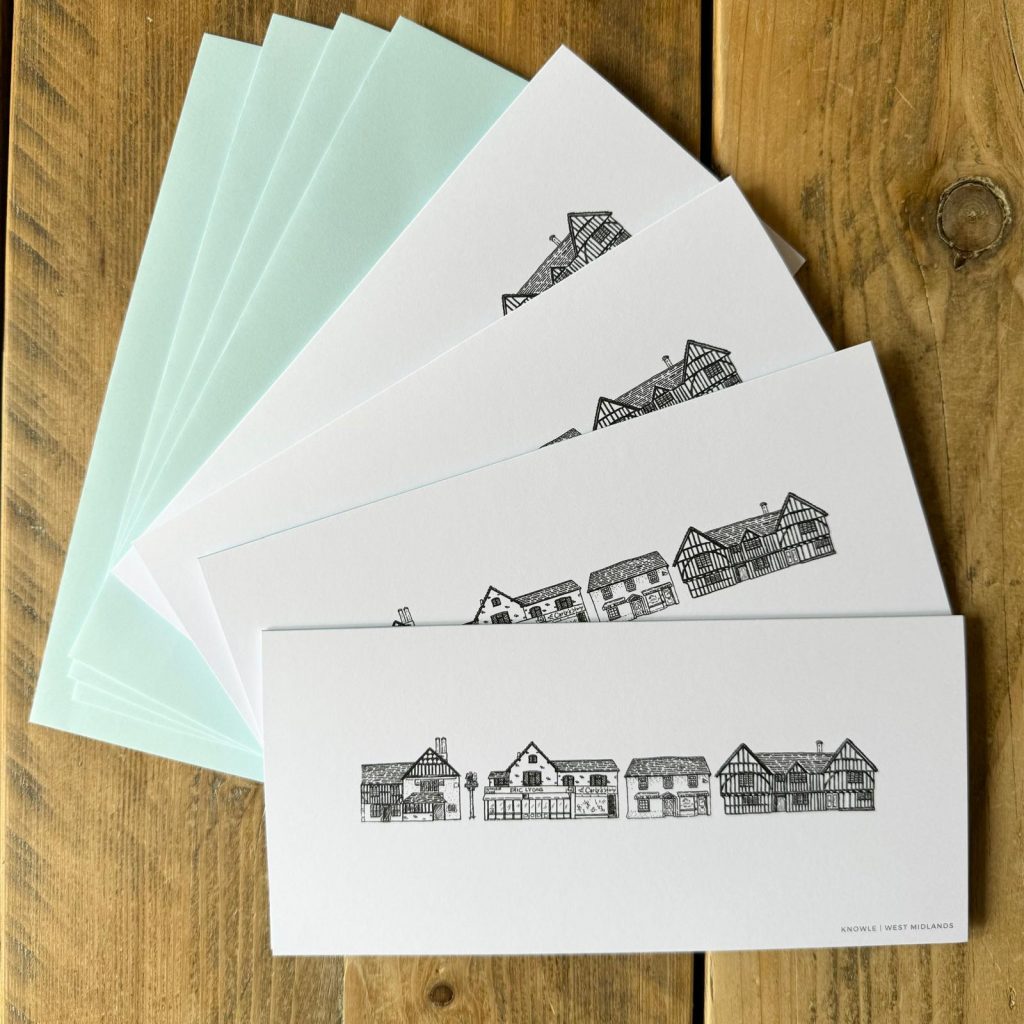 Four cards on a wooden table. The identical cards are white with black line drawing of distinctive buildings in Knowle