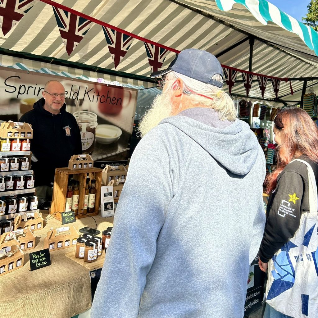 Shoopers browse a stall at a street market. The market trader is talking to a customer.