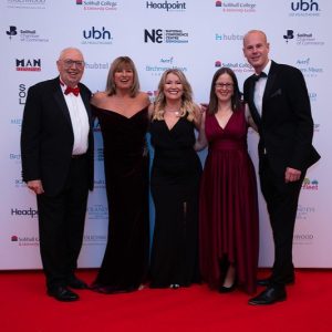 Some of the Visit Knowle team stand in front of of an advertising board at an awards ceremony. They are all in smart dress, arms linked around each other smiling to the camera