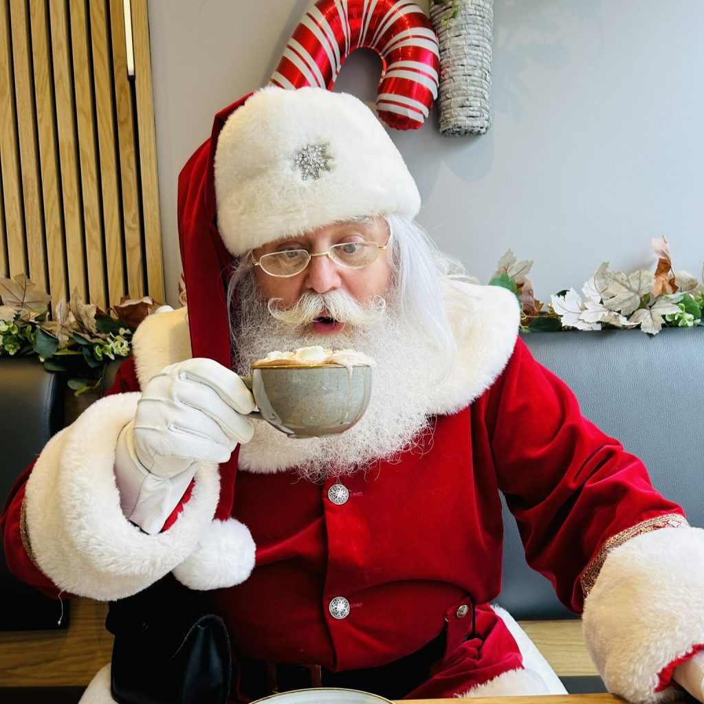 Santa drinking hot chocolate in a cafe with a red candy cane behind him and a cake on the table