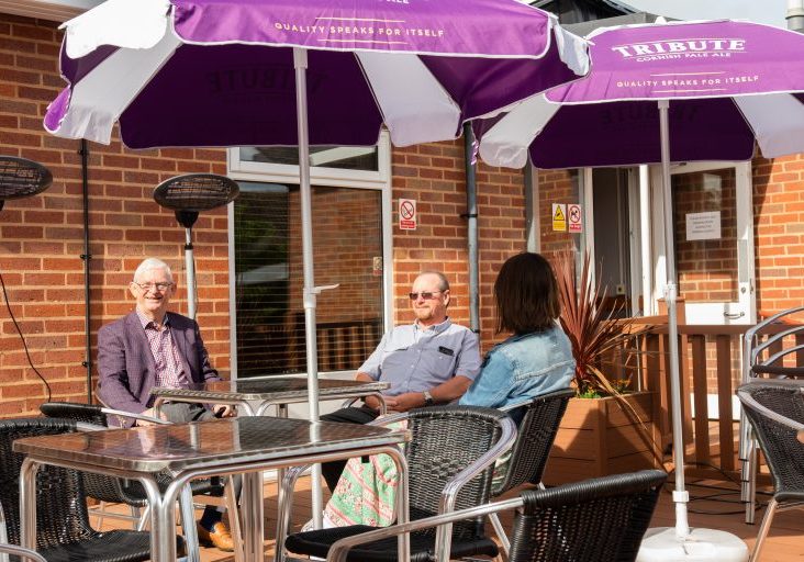 Outdoor seating at Royal British Legion Ken of Knowle British Legion sits with a colleague and a customer at a table with a sun umbrella outside Knowle British Legion